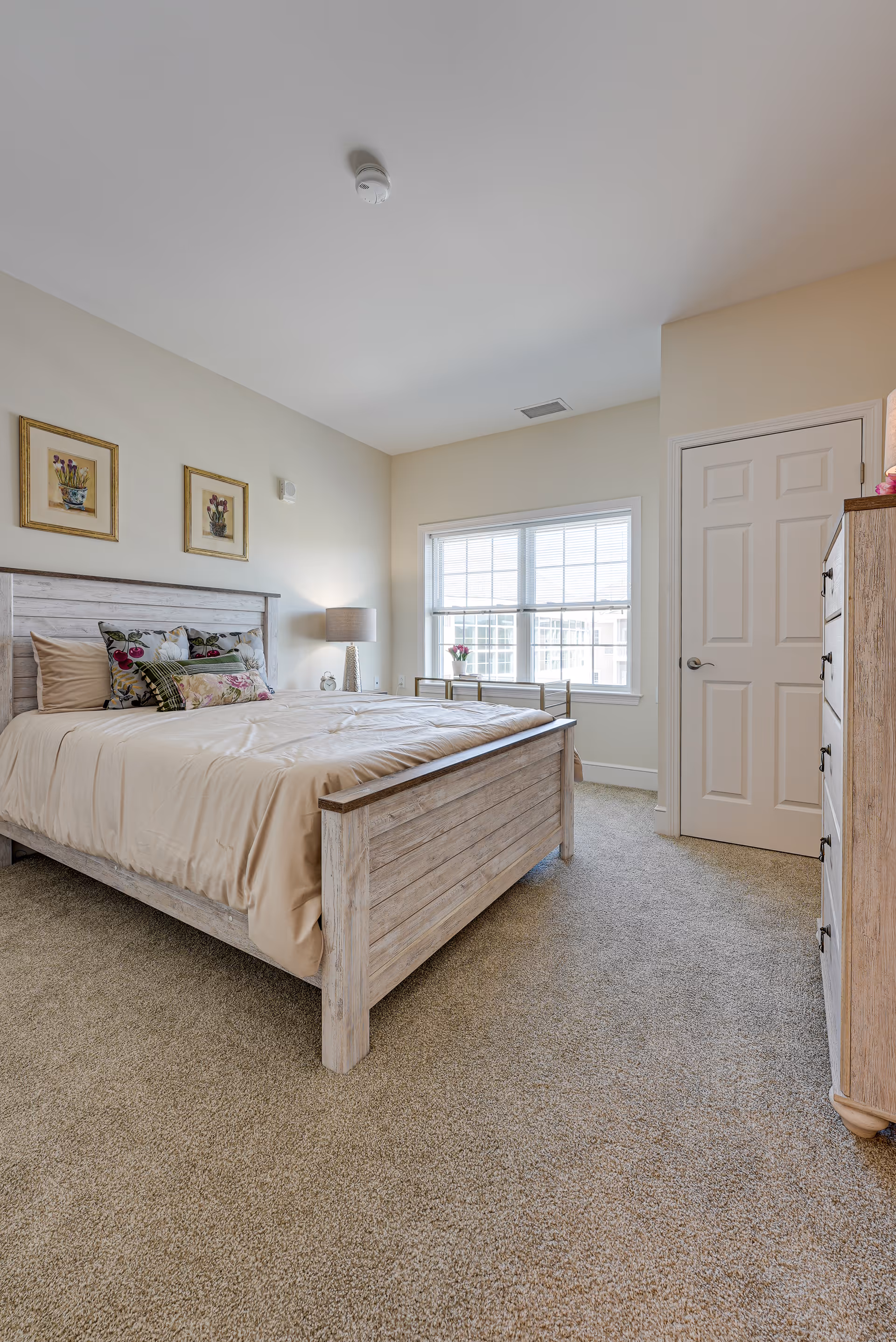 A bright bedroom with a wooden bed frame and beige bedding, decorated with floral pillows. There are two framed floral pictures on the wall above the bed. A bedside table with a lamp is next to the bed. A window with blinds lets in natural light, and a wooden dresser is positioned near a closed white door. The room has beige carpet flooring and light-colored walls.