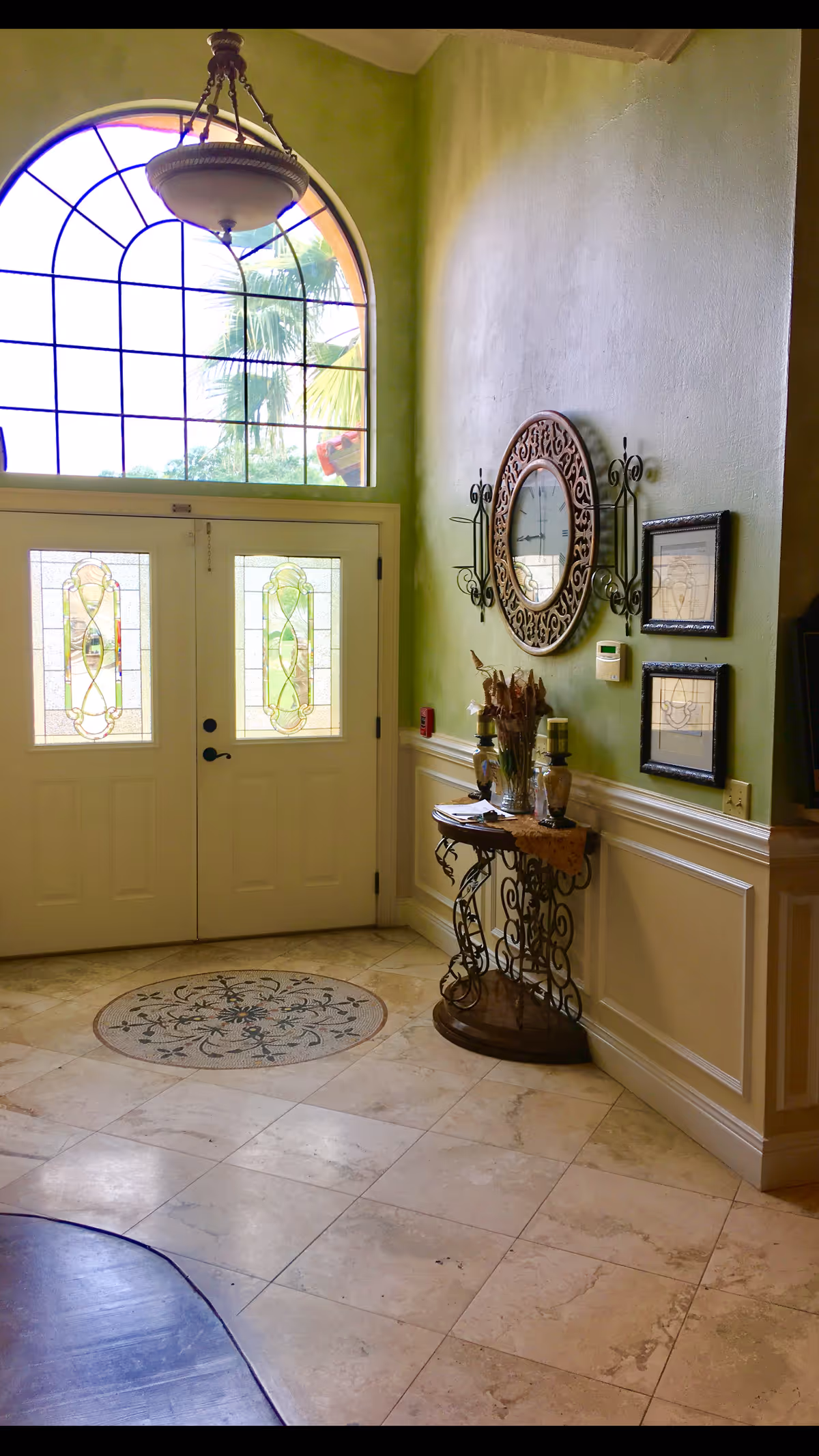 Entrance area with double doors featuring decorative glass panels, a large arched window above letting in natural light, a hanging chandelier, and a small ornate table with decorative items against a light green wall with a round mirror and framed pictures.