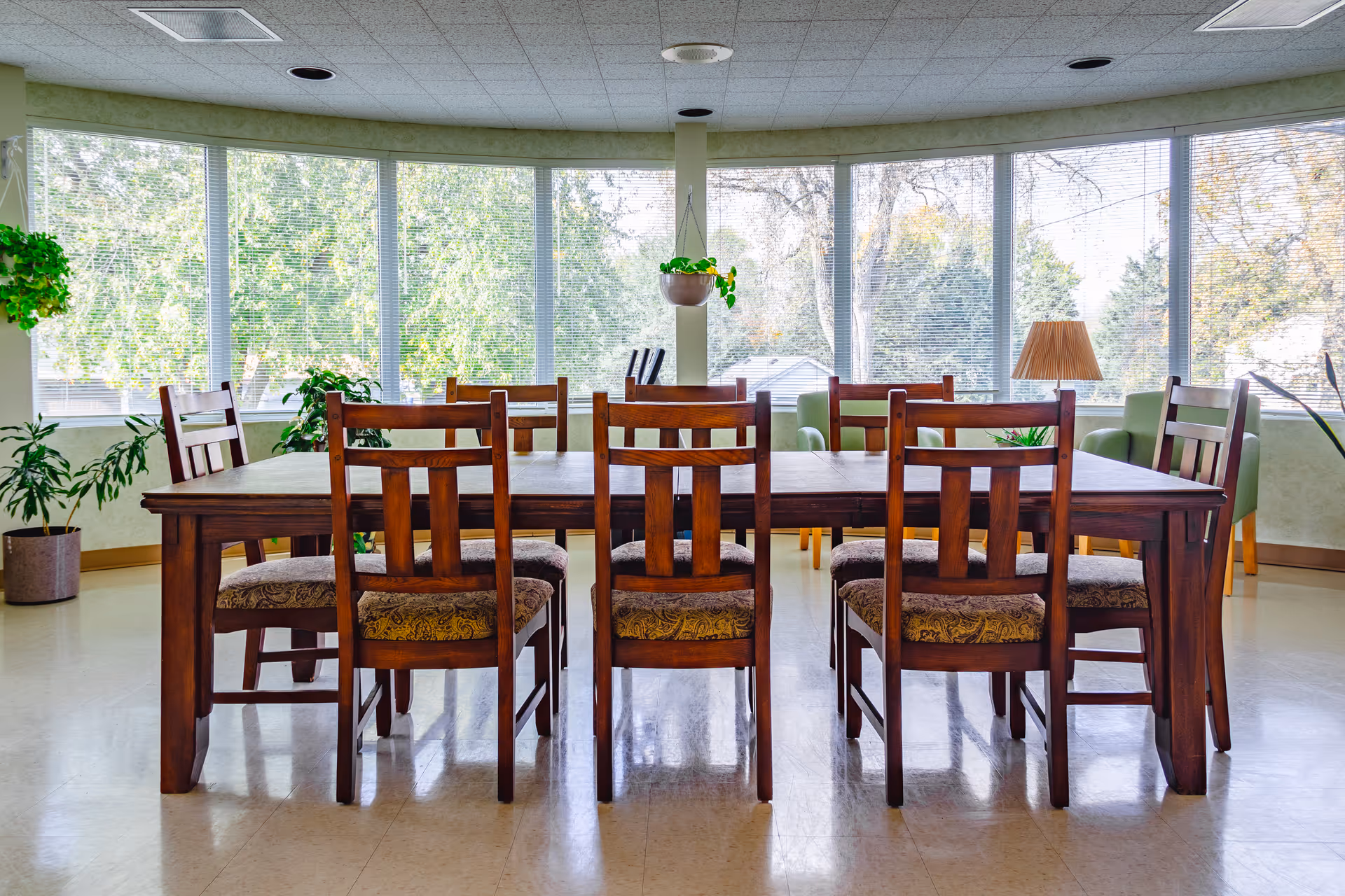 Wooden dining table with eight chairs in a bright common room with large bay windows and potted plants.