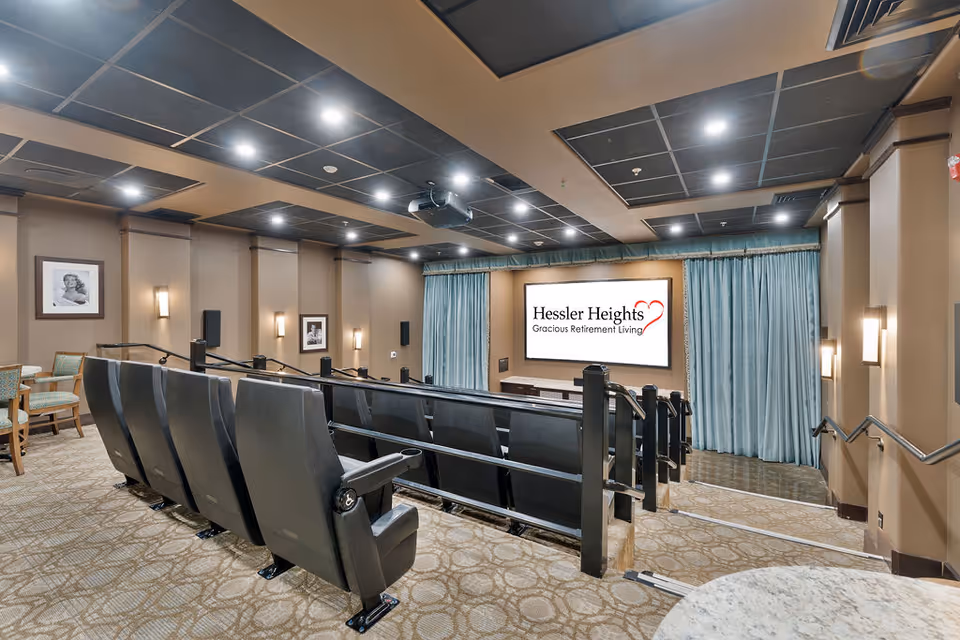 A small theater room with tiered seating featuring black cushioned chairs. The room has beige walls with framed black and white photos, wall sconces, and a ceiling with recessed lighting. At the front, there is a large screen displaying the text 'Hessler Heights Gracious Retirement Living' with a blue curtain backdrop. Handrails are present along the steps leading down to the screen.