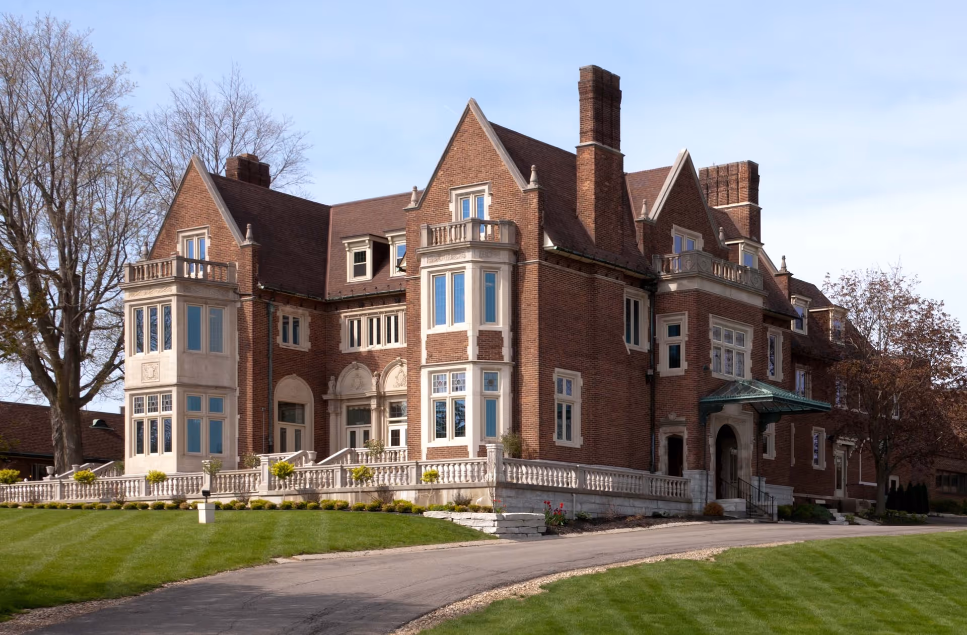 Large brick building with multiple chimneys and large windows, surrounded by a well-maintained lawn and trees under a clear sky.