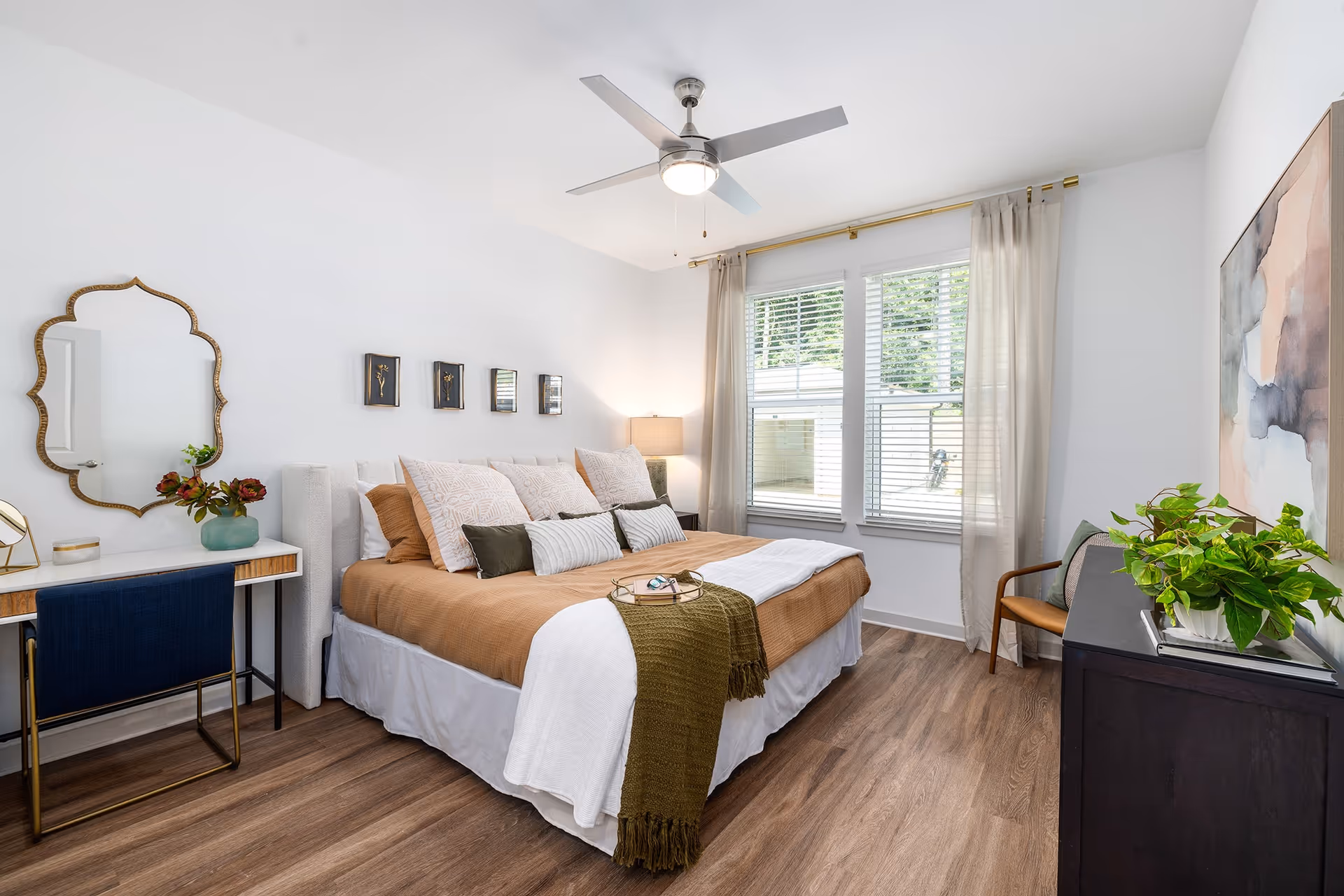 A bright and modern bedroom with a large bed featuring brown and white bedding and multiple pillows. There is a ceiling fan with a light above the bed. To the left, a decorative mirror hangs above a small desk with a blue chair and a vase with flowers. On the right side, a dark wooden dresser holds a green potted plant and a large abstract painting hangs above it. Two windows with white blinds and sheer curtains allow natural light into the room. The floor is wood with a warm tone.