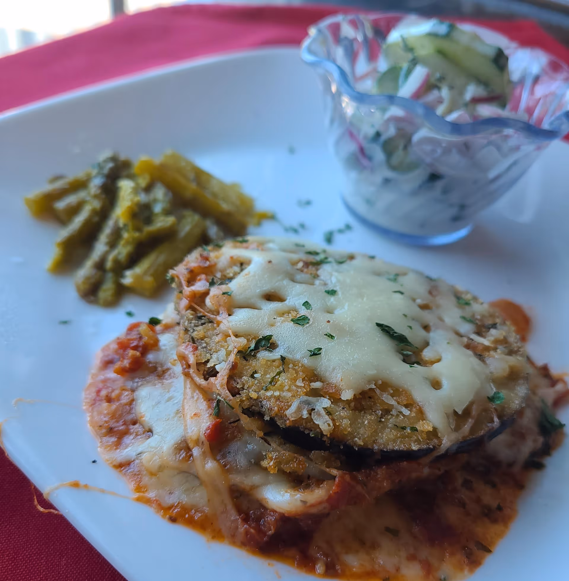 Close-up of a plated baked breaded eggplant topped with melted cheese, served with green beans and a small cucumber-radish salad in a glass bowl on a white plate.
