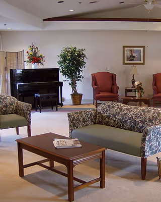 Communal living room with patterned sofas, armchairs, a wooden coffee table, a piano, and potted plants.