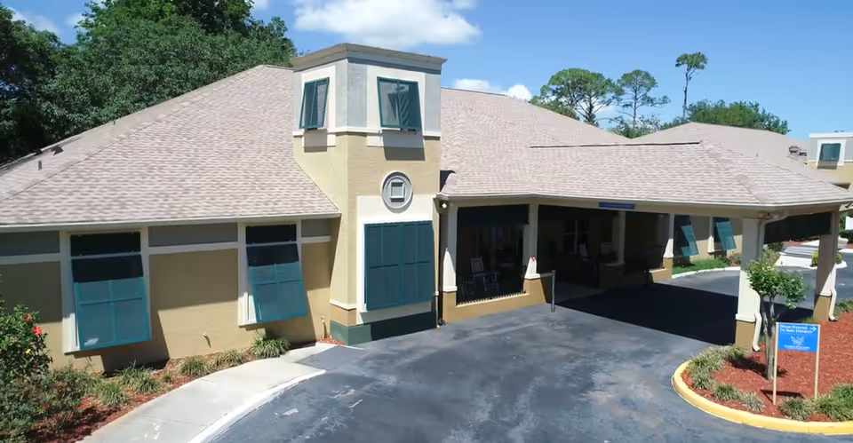 Front exterior of a memory care facility showing a covered entrance with a porte-cochere, teal window shutters, driveway and landscaping.