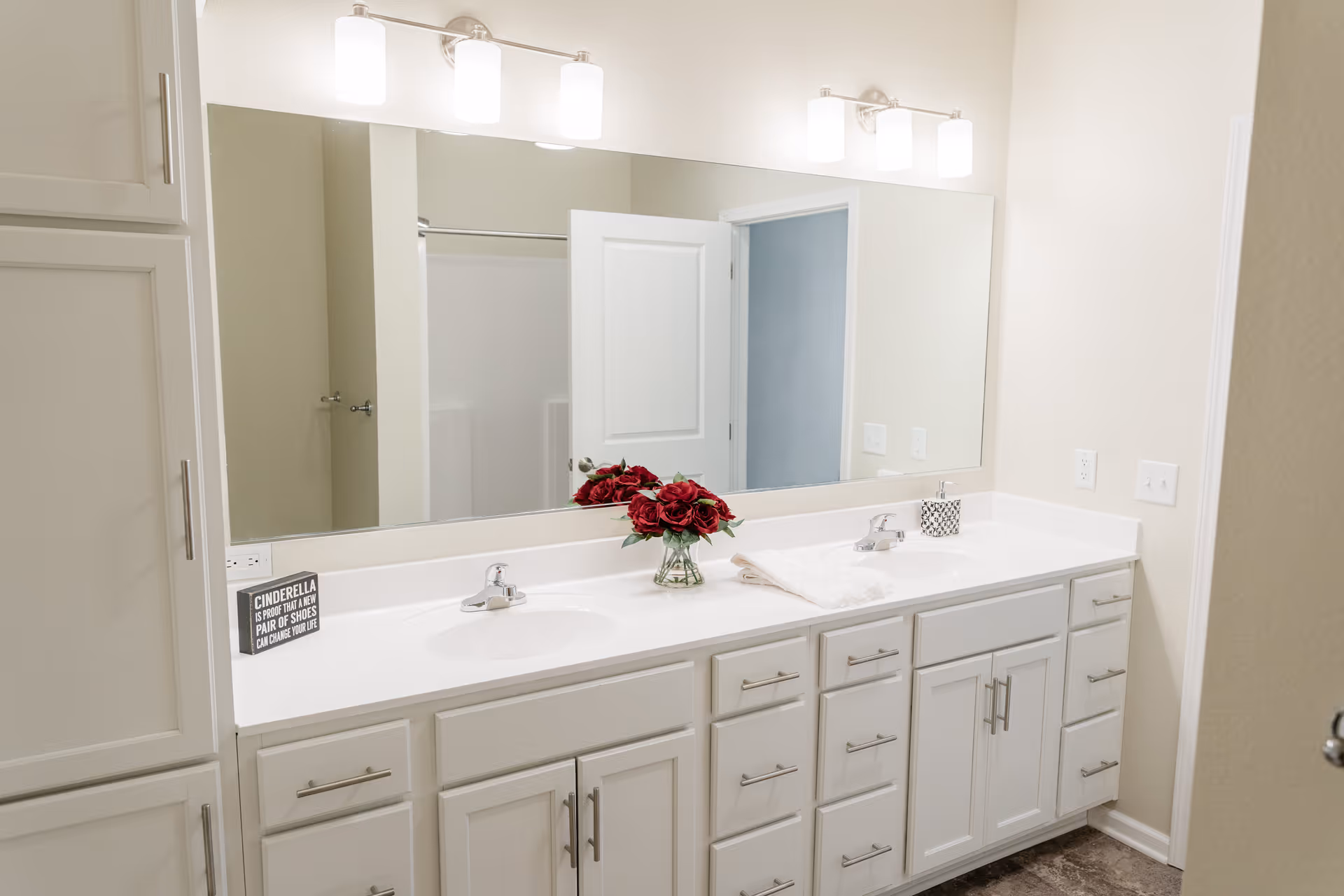 A clean, modern bathroom vanity with two sinks, white cabinetry, and a large mirror above. There are two sets of three-light fixtures mounted above the mirror. On the countertop, there is a small vase with red flowers, a folded white towel, a soap dispenser, and a small decorative sign. The background shows a partially open door revealing a shower area.