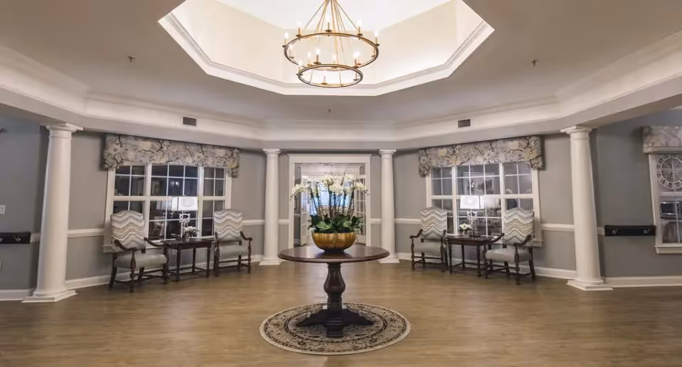A spacious, elegant common area with a round wooden table in the center holding a large floral arrangement. The room features a high, octagonal ceiling with a chandelier, two sets of windows with patterned valances, and pairs of cushioned chairs with small tables and lamps beside them. The walls are painted gray with white trim and columns, and the floor is wood with a small round rug under the table.