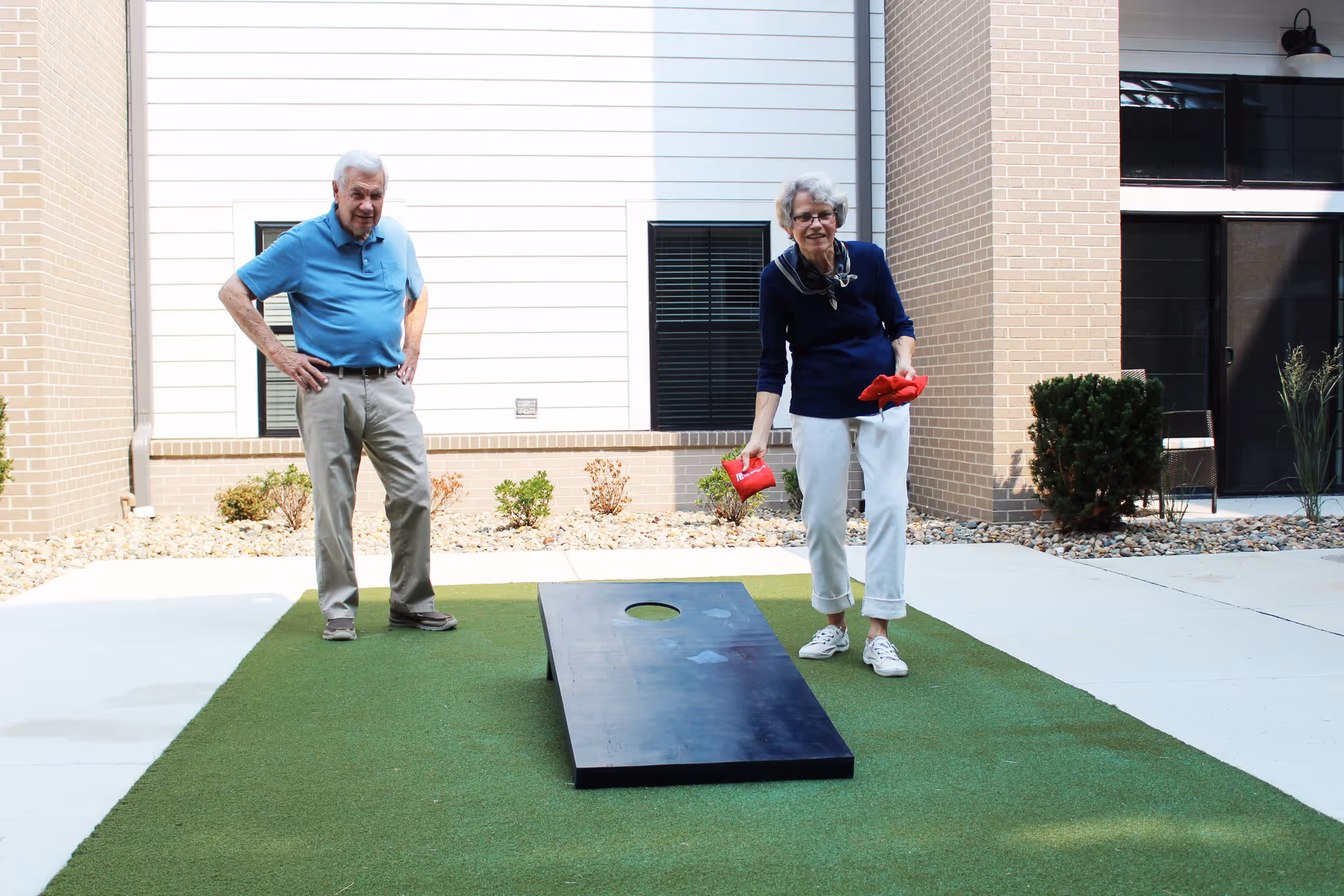 An elderly man and woman playing cornhole on a green artificial turf area outside a building. The woman is tossing a red bean bag towards a black cornhole board with a hole, while the man watches with his hands on his hips. The background shows a brick and white siding exterior with windows and some landscaping.