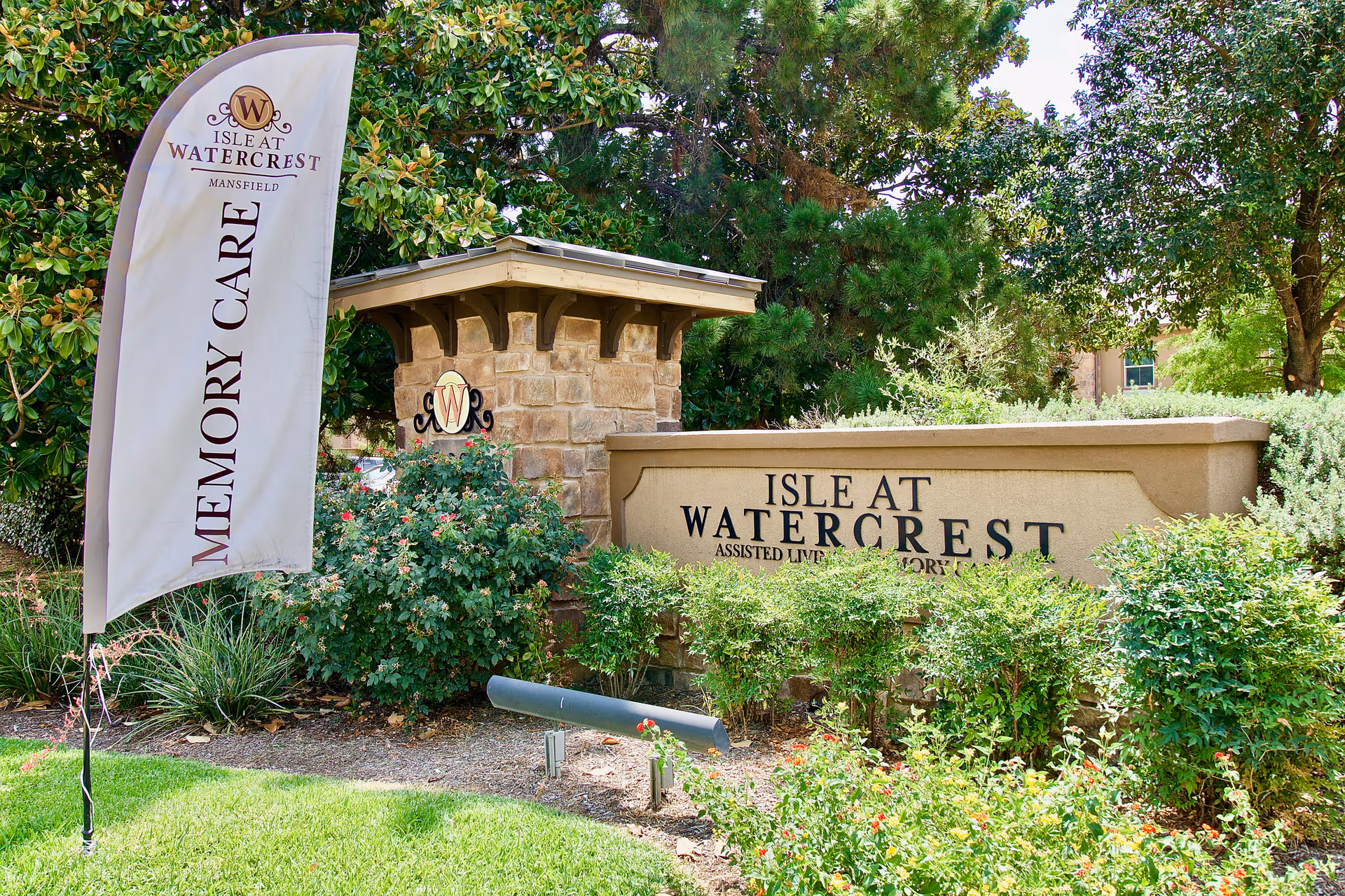 Outdoor view of the Isle at Watercrest Mansfield assisted living and memory care facility sign surrounded by greenery and trees, with a flag displaying 'Memory Care'.