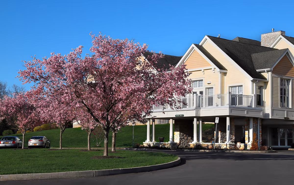 Exterior view of a senior living facility building with beige siding and white trim, featuring a covered entrance supported by columns. In the foreground, there are several blooming pink cherry blossom trees and a green lawn, with a clear blue sky above. Two cars are parked along the driveway near the trees.