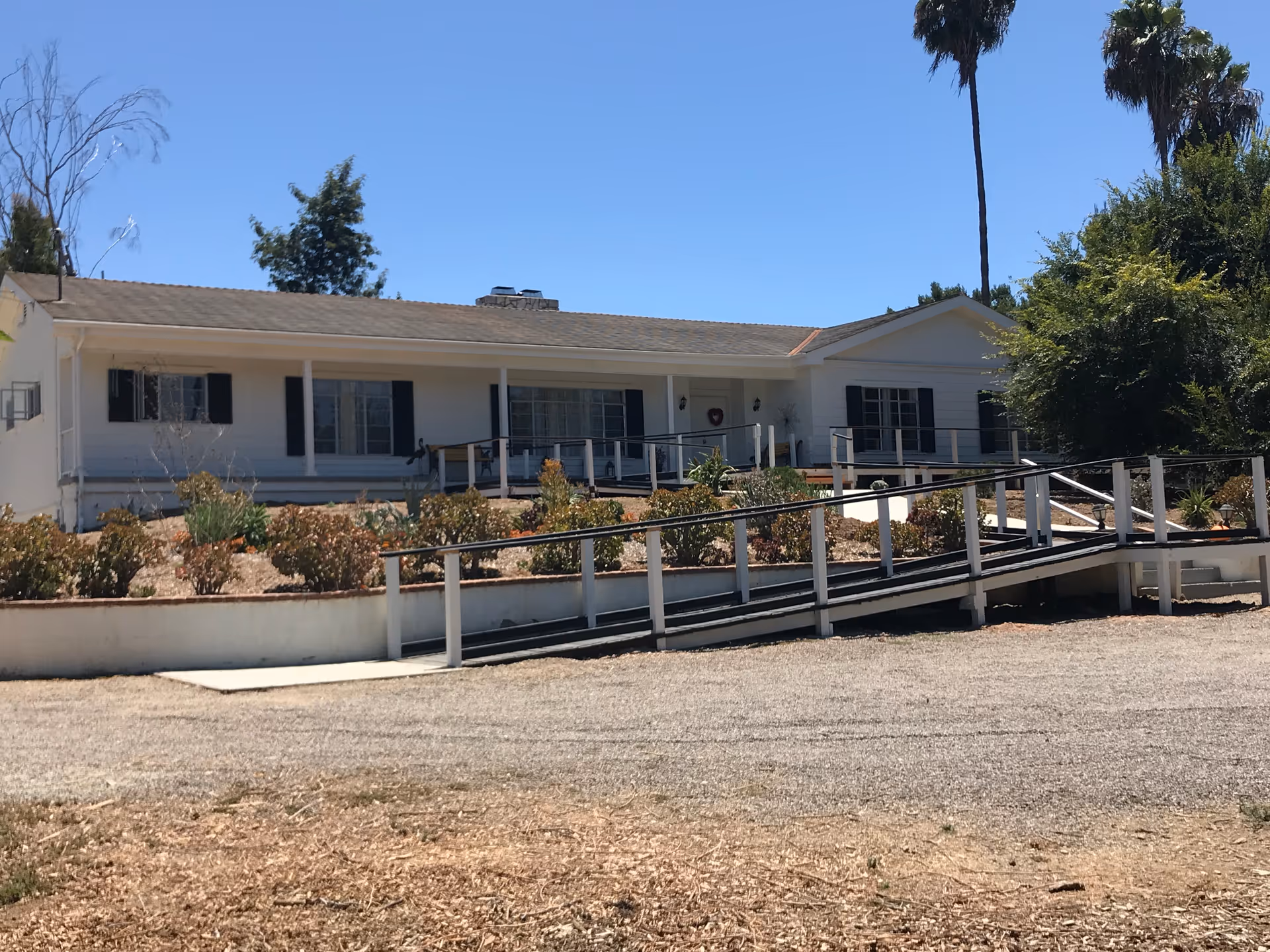 Single-story white senior living cottage with a ramped front porch, landscaping, and palm trees under a clear blue sky.