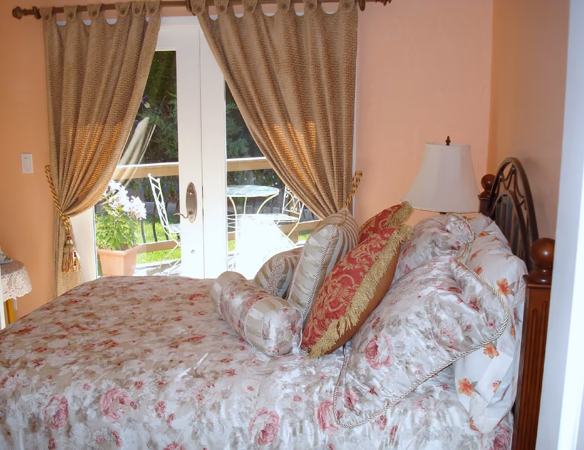A bedroom with a floral-patterned bedspread and multiple decorative pillows. The bed has a wooden headboard with metal detailing. Next to the bed is a white lampshade on a wooden nightstand. Behind the bed, there are glass double doors with beige curtains tied back, leading to an outdoor patio with a metal table and chairs and some potted plants.