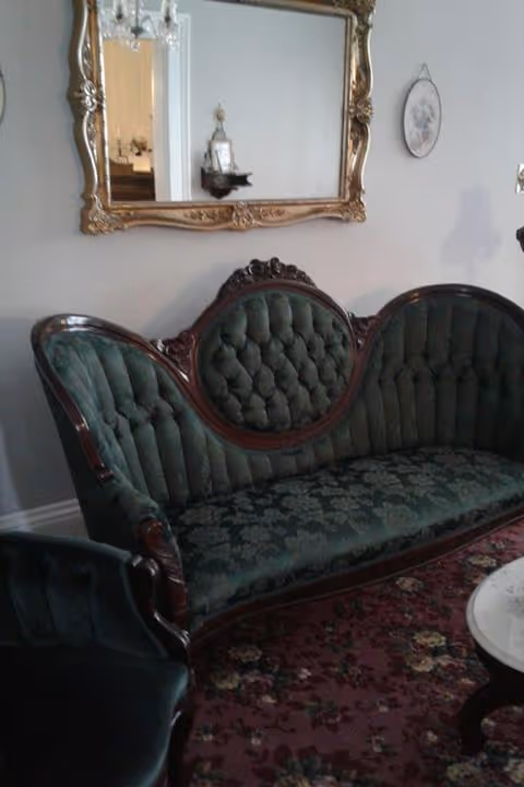 An antique-style green velvet tufted sofa with wooden frame sits against a light gray wall. Above the sofa is an ornate gold-framed mirror reflecting part of the room. To the right of the mirror is a small oval wall decoration. A floral patterned carpet covers the floor, and a small round marble-top table is partially visible in the foreground.