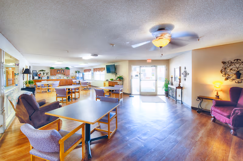 A spacious senior living common area with wooden flooring, multiple tables and chairs, a ceiling fan with light, a large flat-screen TV mounted on the wall, and a kitchen area with wooden cabinets in the background. The room is well-lit with natural light coming through glass doors and windows. There are decorative elements including plants, wall art, and a lamp on a side table.