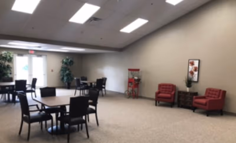 A spacious room with several square tables and black chairs arranged for seating. There are two red armchairs with a small dark wooden cabinet between them, decorated with a plant and a framed floral picture on the wall. A red popcorn machine is positioned near the wall. The room has beige walls, carpeted floor, and fluorescent ceiling lights. Double glass doors and potted plants are visible in the background.