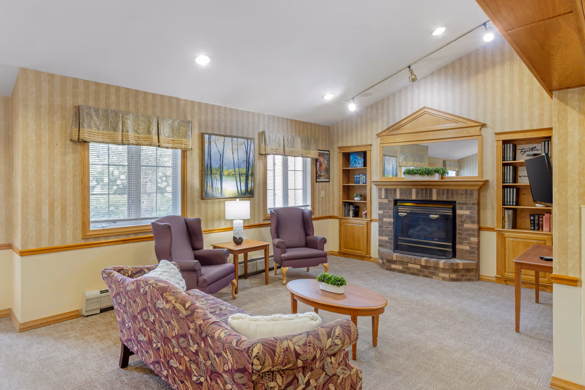 A cozy living room with beige striped wallpaper and carpeted floor. The room features a floral-patterned sofa with white pillows, two purple armchairs, a wooden coffee table with a small plant, and a side table with a lamp. There is a brick fireplace with a wooden mantel and a large mirror above it. Built-in wooden shelves flank the fireplace, holding books and decorative items. Two windows with blinds and valances let in natural light, and framed artwork hangs on the walls.