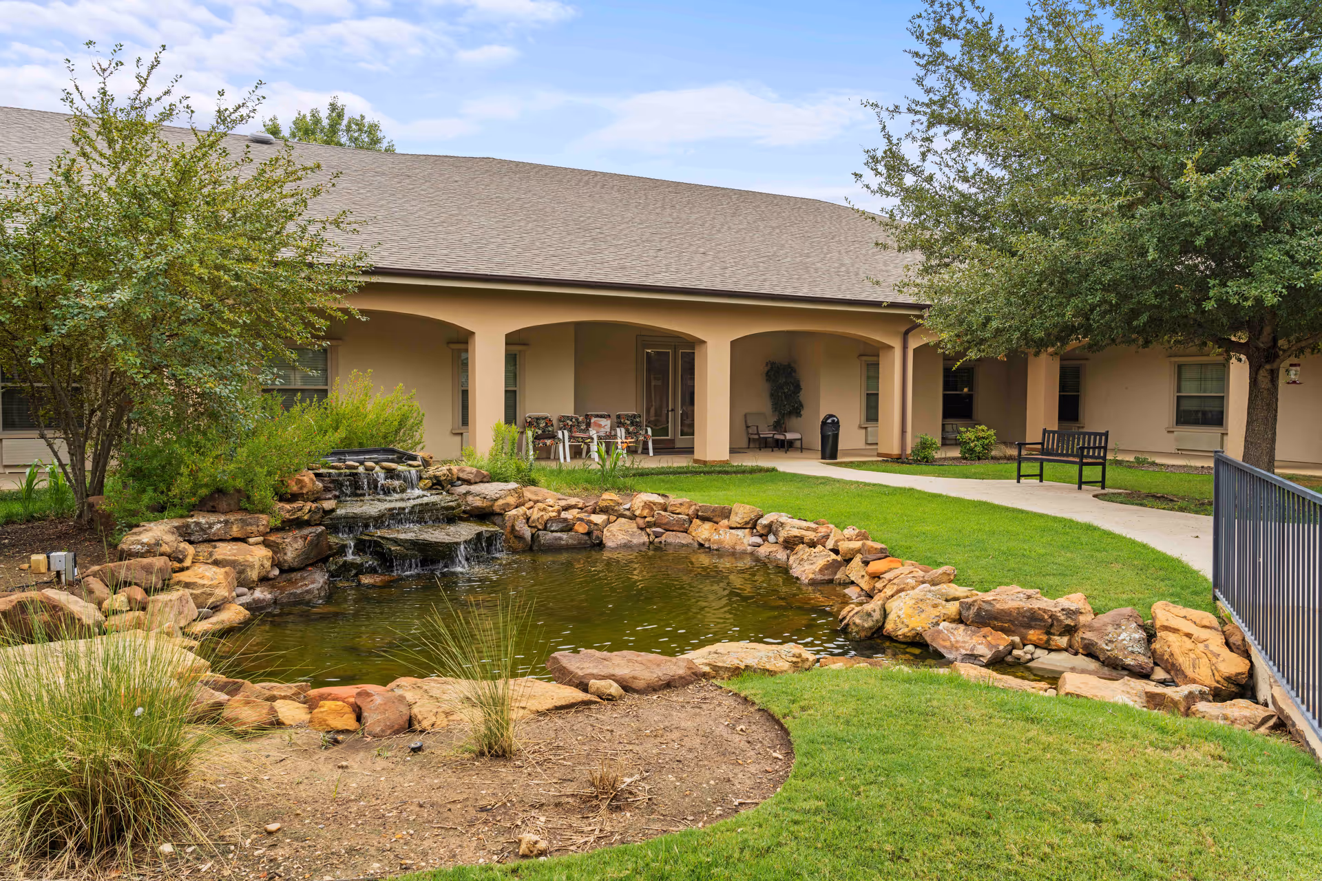 Outdoor garden area at Volante Senior Living of Grapevine featuring a small pond with a rock waterfall, surrounded by green grass, trees, and shrubs. There is a paved walkway, benches, and a covered patio area with chairs in the background.