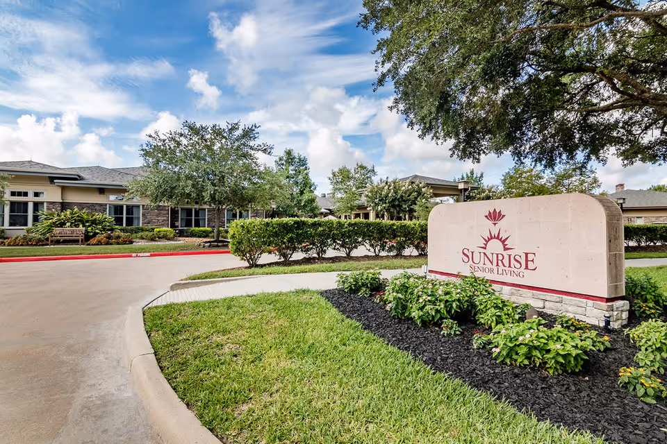 Exterior view of Sunrise Senior Living facility with a large stone sign displaying the name and logo. The building is surrounded by green bushes, trees, and a well-maintained lawn under a partly cloudy sky.