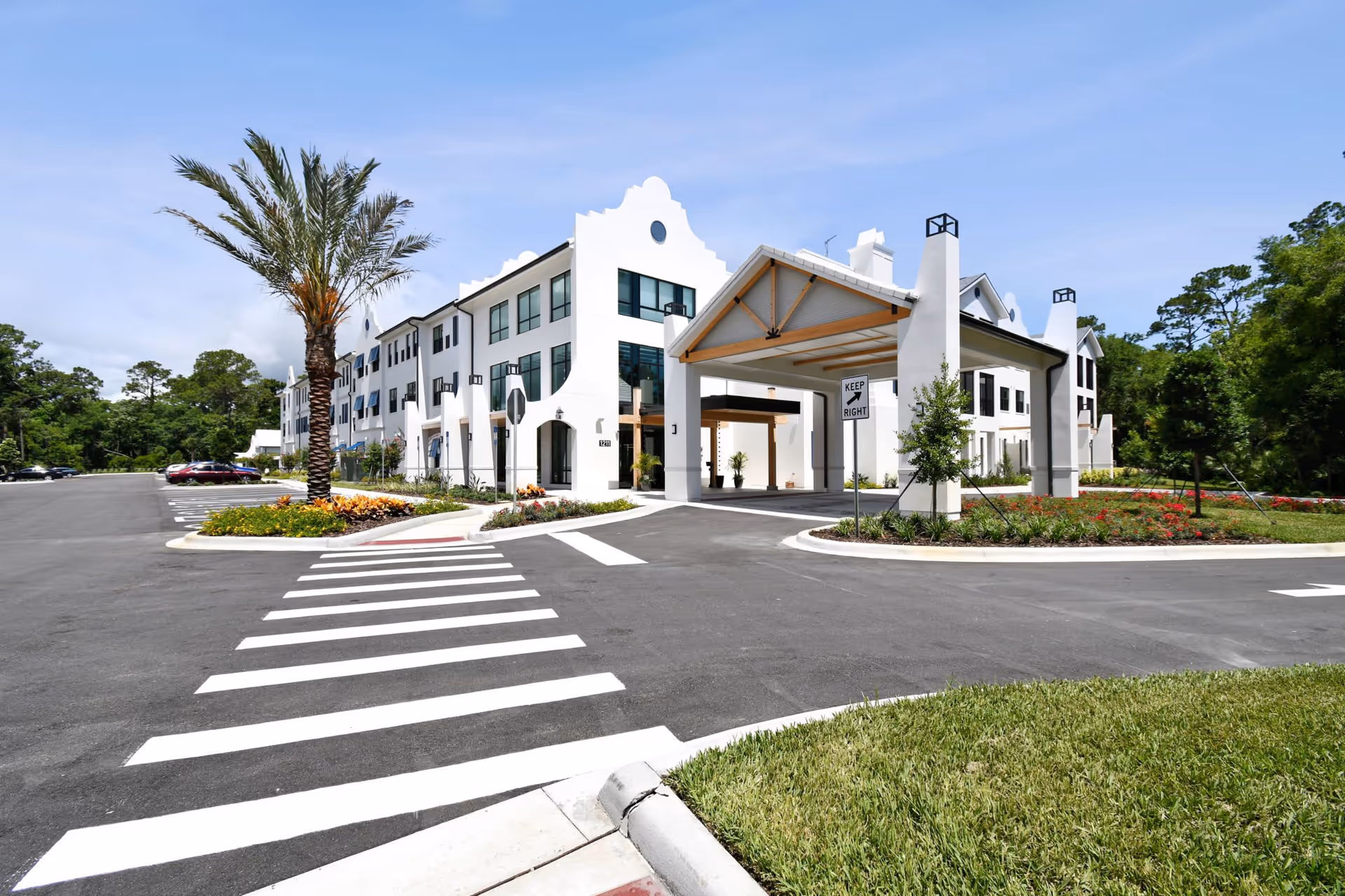 Exterior view of a modern senior living facility with white walls and large windows, featuring a covered entrance with a 'Keep Right' sign, landscaped flower beds, palm trees, and a parking lot under a clear blue sky.