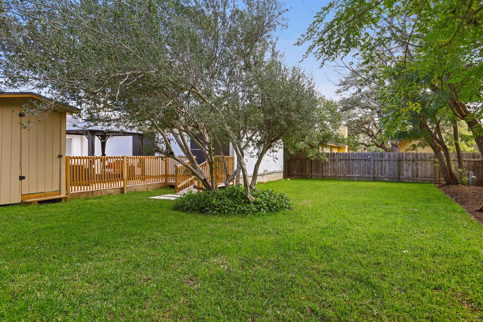 A fenced backyard with green grass, a small tree in the center surrounded by plants, a wooden shed on the left, and a wooden fence enclosing the area. Trees and a clear blue sky are visible in the background.