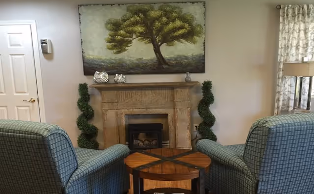 A cozy living room area with two green plaid armchairs facing a wooden coffee table and a stone fireplace. Above the fireplace is a large painting of a tree. On either side of the fireplace are two spiral-shaped topiary plants. To the right, there is a window with patterned curtains and a floor lamp.
