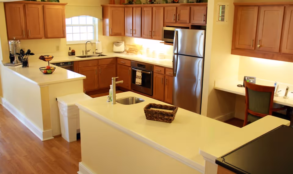 A bright and spacious kitchen with wooden cabinets, a stainless steel refrigerator, oven, and microwave. The kitchen features a white countertop island with a small sink and a wicker basket. There is a window above the main sink, and a small desk area with a chair is visible in the corner.