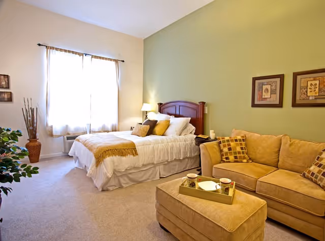 A cozy bedroom with a large bed featuring white bedding and multiple pillows, a wooden headboard, and a bedside table with a lamp. Next to the bed is a beige sofa with patterned cushions and a matching ottoman with a tray holding two cups and a plate. The room has a green accent wall with two framed pictures and a window with sheer curtains letting in natural light.