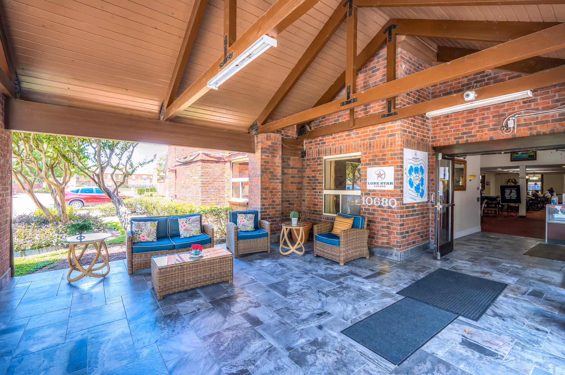 Covered outdoor seating area at Lone Star Living facility with wicker furniture including a sofa, two chairs, and a coffee table, all with blue cushions and floral pillows. The area has a tiled floor, wooden ceiling beams, and brick walls. There is a sign on the wall with the Lone Star Living logo and the number 10680. The entrance to the building is visible with open double doors leading inside.