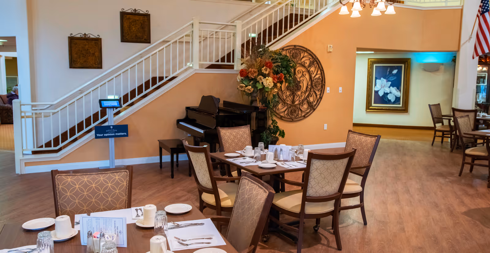 Dining area in Holiday Lone Oak facility with tables set for meals, chairs with patterned upholstery, a black piano against a peach-colored wall, decorative floral arrangement, and a staircase with white railing in the background.