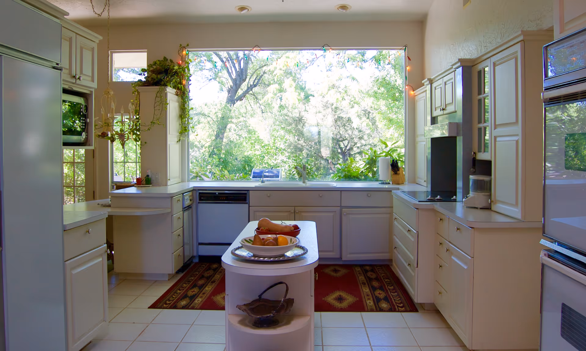 Bright kitchen with white cabinets and appliances, a small island with fruit bowls, and a large window overlooking lush green trees outside. The kitchen has a tiled floor and two patterned rugs in front of the sink and stove areas.