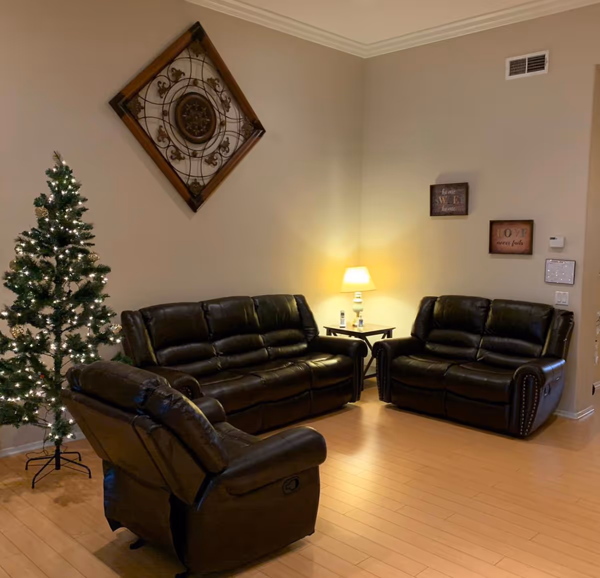 A cozy living room with three dark leather recliner sofas arranged around a corner. A small side table with a lit lamp and some items is placed between two sofas. A decorated Christmas tree with white lights stands in the corner. The walls are beige with decorative wall art, including a large square metal and wood piece and two smaller framed quotes.