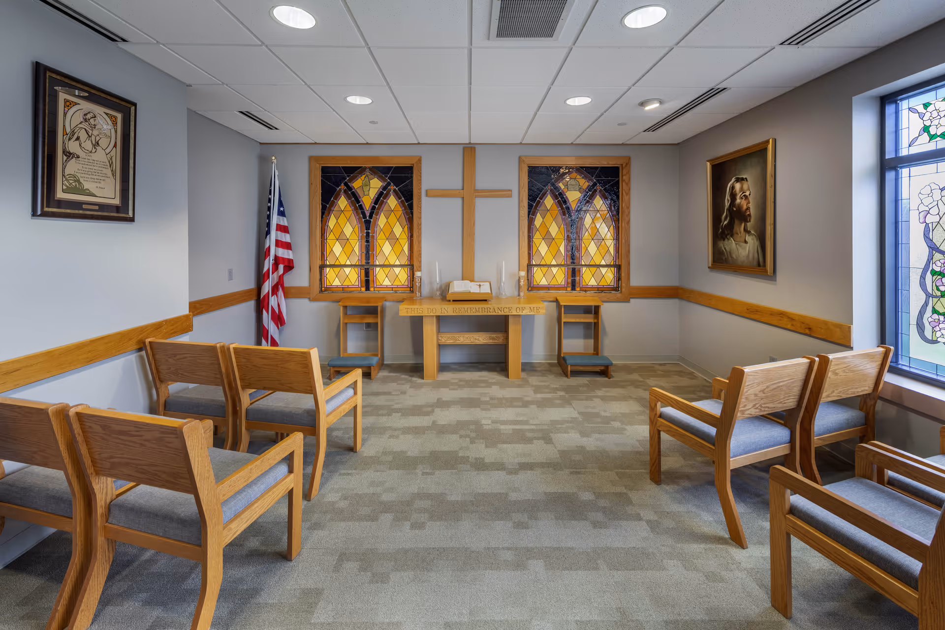 A small chapel room with wooden chairs arranged in rows facing a wooden altar with a cross mounted on the wall behind it. The altar has two stained glass windows on either side and an open Bible on top. An American flag stands in the corner, and there are framed religious pictures on the walls. The room has a carpeted floor and a ceiling with recessed lighting.