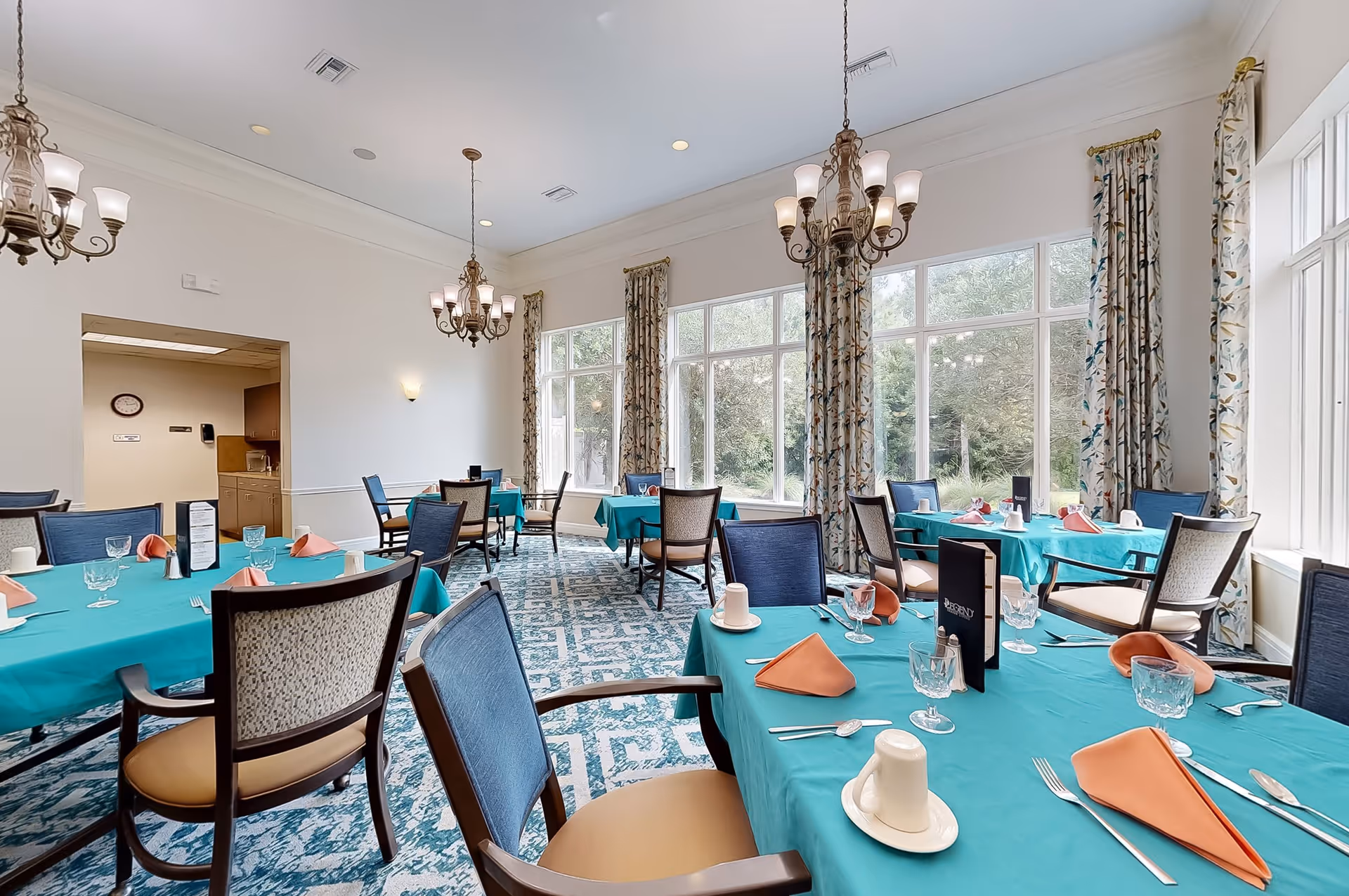 A bright dining room with large windows letting in natural light, decorated with patterned curtains. Several tables are covered with teal tablecloths and set with orange napkins, glassware, and white cups. The room features elegant chandeliers hanging from the ceiling and a patterned carpet on the floor.
