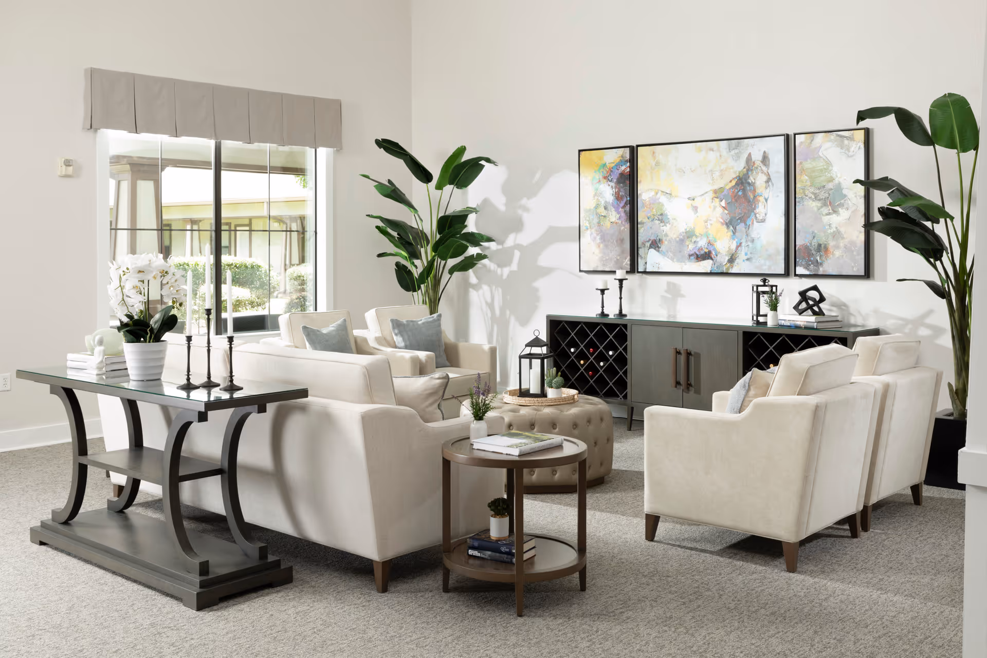 A bright and spacious living room with beige upholstered sofas and armchairs arranged around a round tufted ottoman. There is a dark wooden console table with decorative items including candles and a potted plant. A large window with a beige valance lets in natural light. The room features a dark wooden sideboard with wine storage and decorative items, and a three-panel colorful horse painting above it. Two large green plants add a touch of nature to the neutral-toned room.