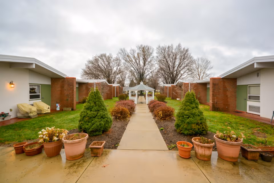 Outdoor walkway lined with small bushes and potted plants leading to a white gazebo, flanked by single-story buildings with brick and white walls under a cloudy sky.