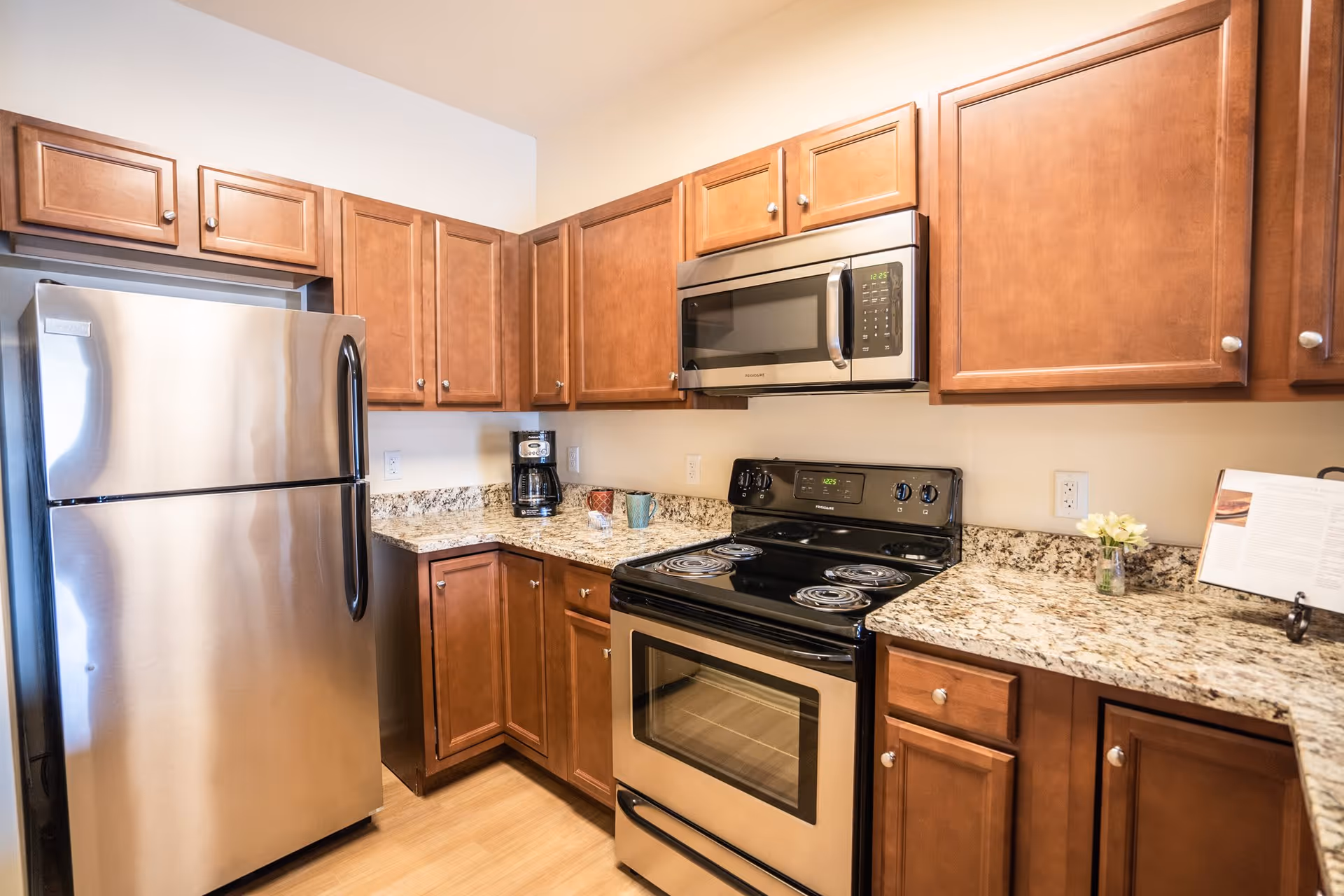 A modern kitchen with wooden cabinets, granite countertops, a stainless steel refrigerator, a black electric stove with oven, a stainless steel microwave above the stove, a coffee maker, and a small vase with flowers on the counter.