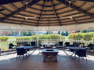 Covered outdoor seating area with a wooden roof structure, several black chairs and benches arranged around a central fire pit, overlooking a green lawn and garden area with bushes and trees in the background.