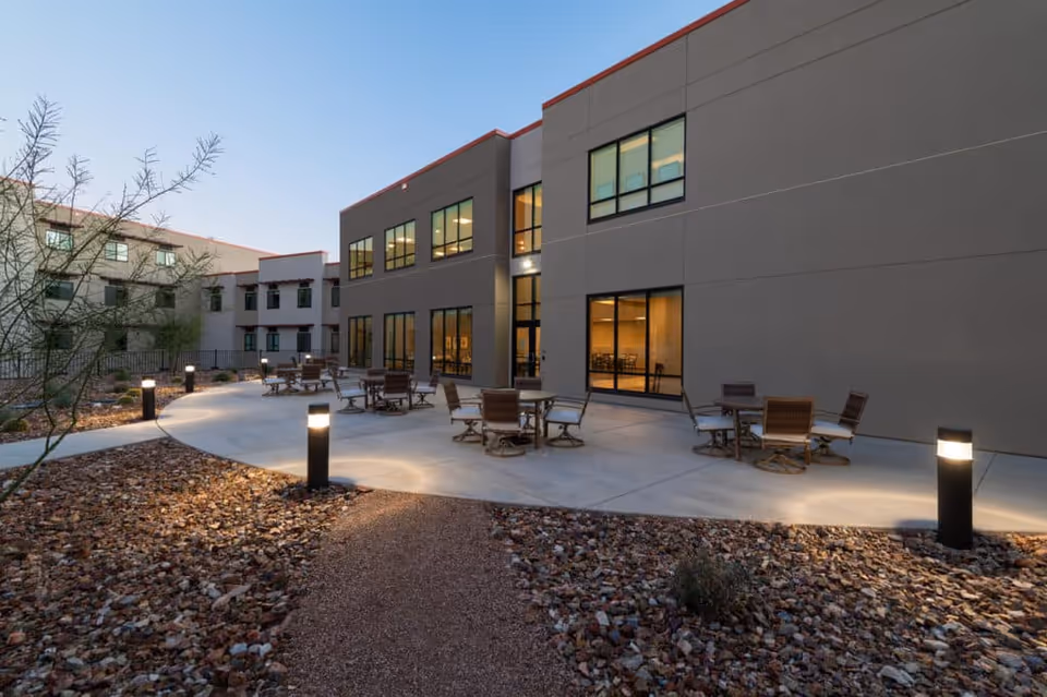 Outdoor patio area at dusk with several round tables and chairs arranged on a concrete surface. The patio is surrounded by a landscaped area with rocks and desert plants. The building adjacent to the patio has large windows with lights on inside.