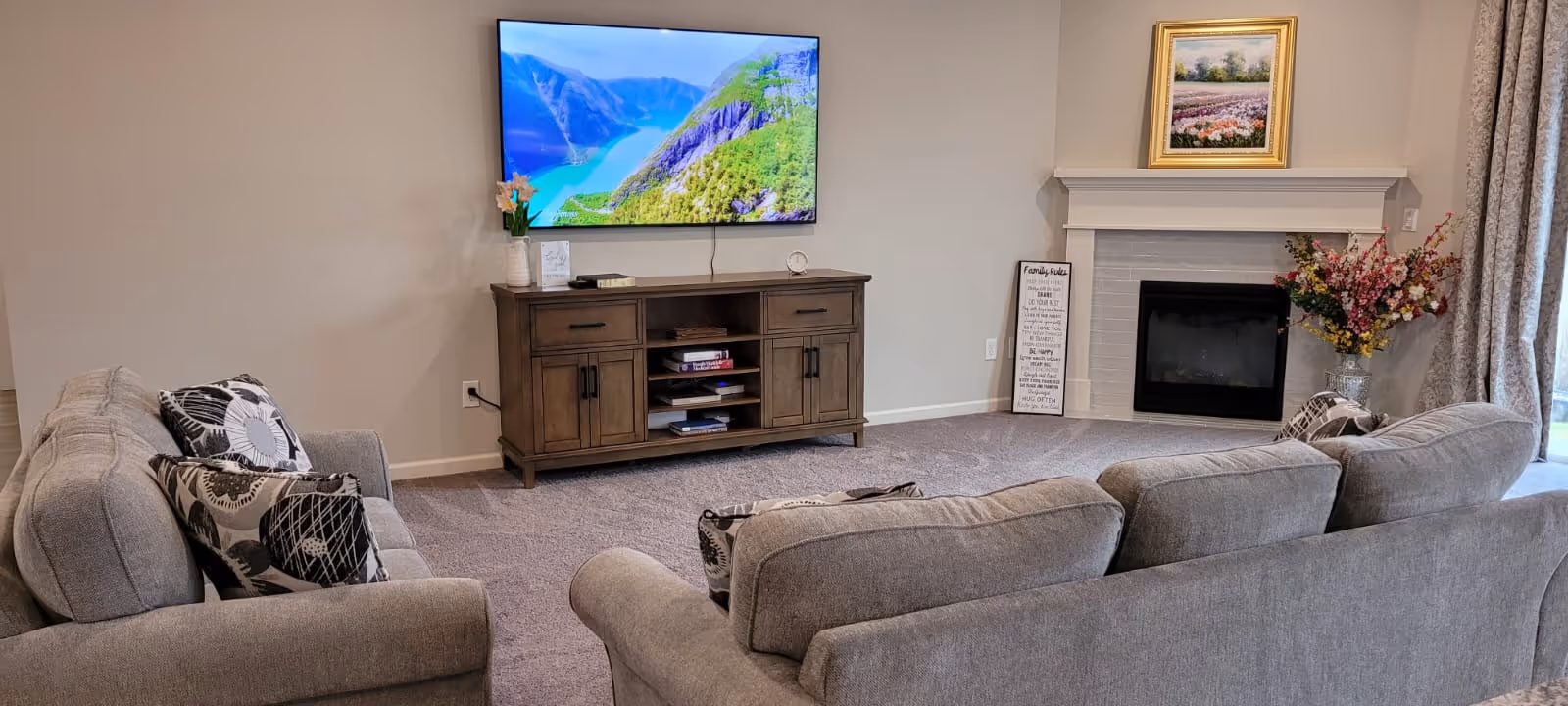 A cozy living room with two gray sofas facing a wall-mounted flat-screen TV displaying a scenic mountain and lake view. Below the TV is a wooden media console with shelves and cabinets. To the right, there is a white fireplace with a framed painting above it and a vase of flowers on the hearth. A decorative sign leans against the wall next to the fireplace, and curtains partially cover a window or door on the far right.