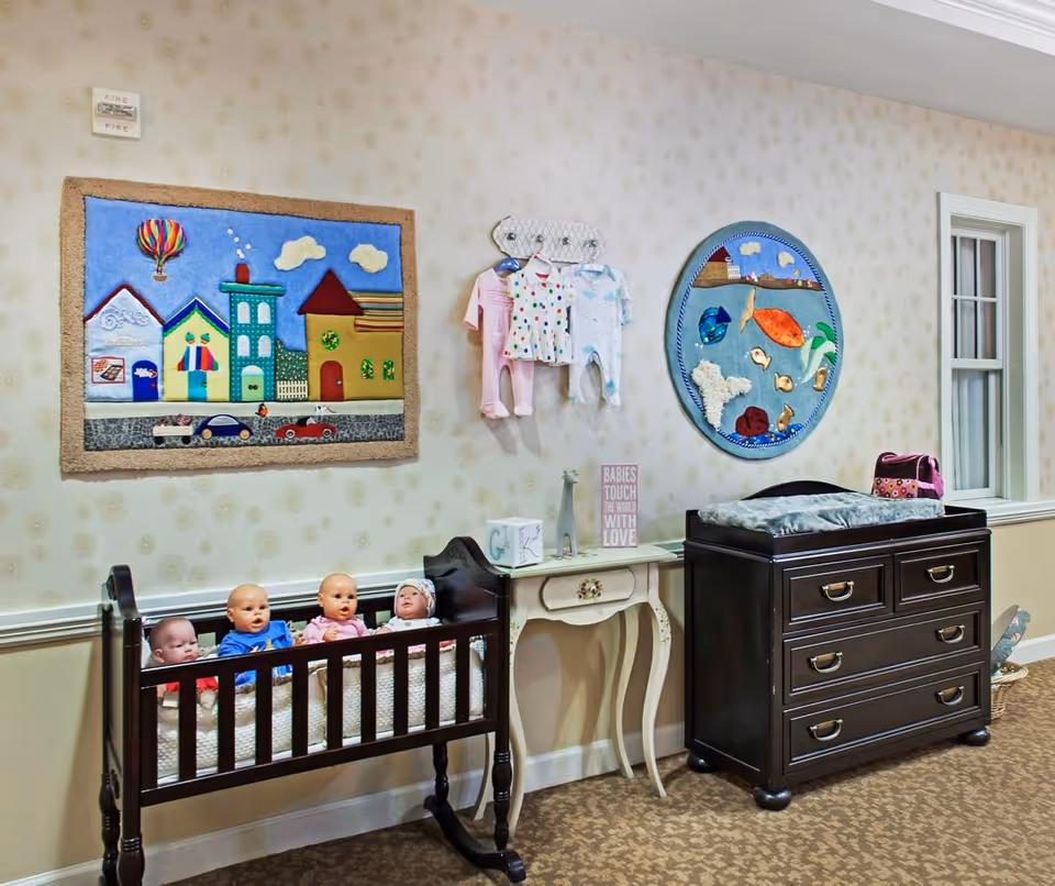 A nursery room with a wooden crib containing four baby dolls, a small white table with decorative items, and a dark wooden changing table with drawers. The walls are decorated with colorful fabric art pieces depicting houses, a hot air balloon, and sea creatures. Baby clothes hang on a rack above the table.