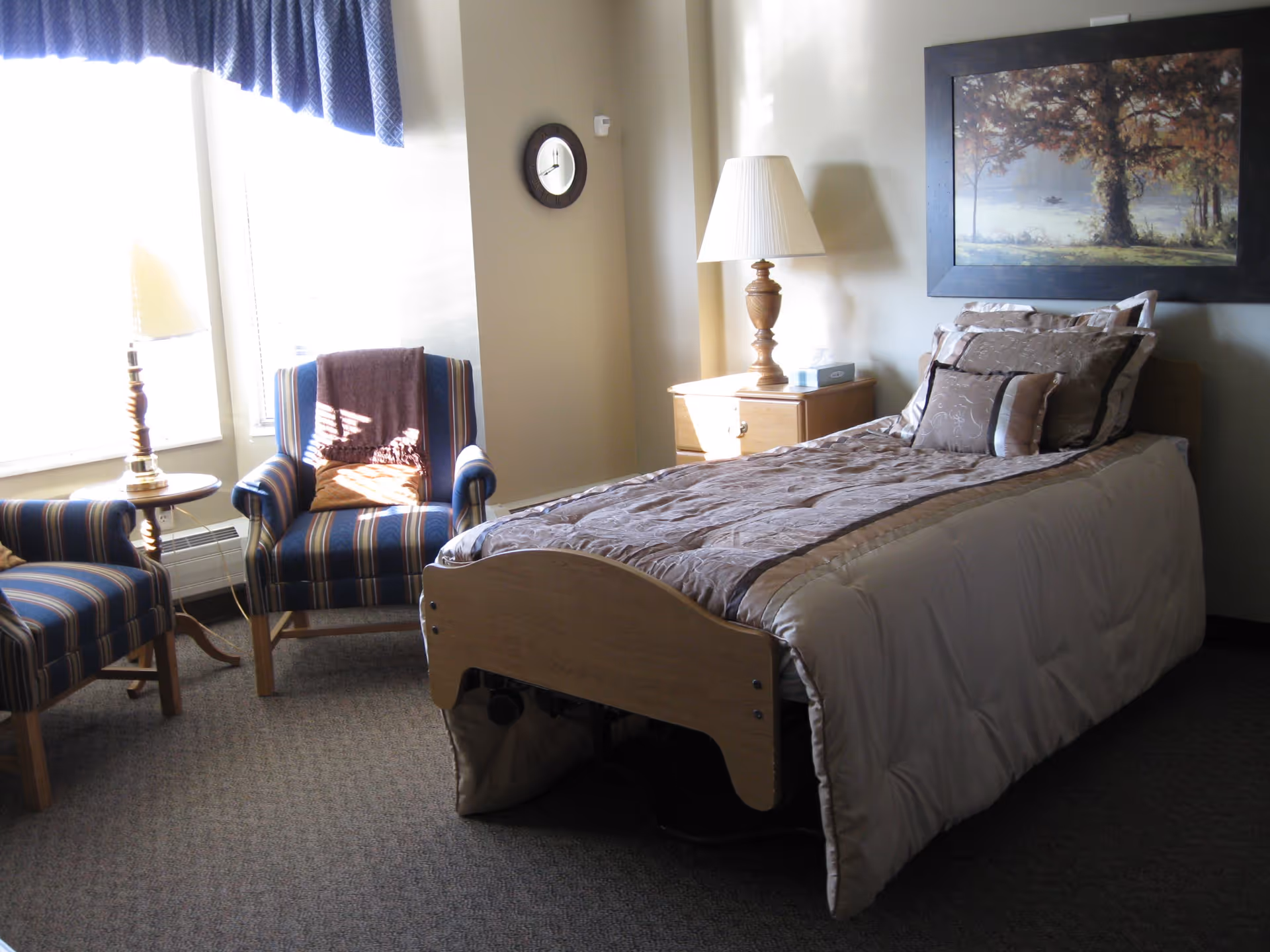 A cozy bedroom with a single bed covered in a beige and brown comforter, a wooden nightstand with a lamp and tissue box, two striped armchairs with a small round table and lamp between them, a wall clock, and a framed painting of a tree by a lake.