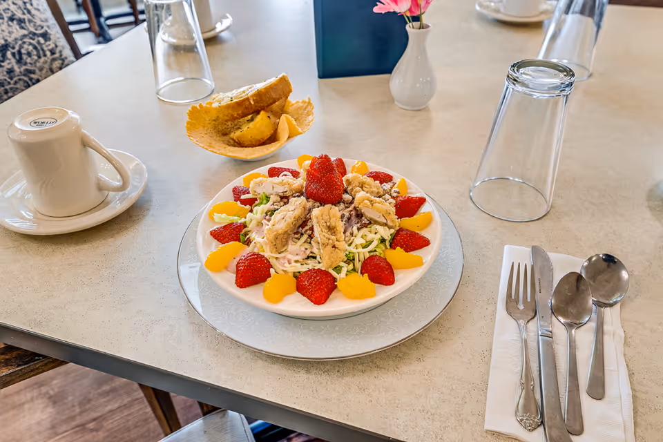 A dining table set with a plate of salad topped with fried chicken strips, strawberries, and mandarin orange slices. There is a cup and saucer, an upside-down glass, a small vase with a pink flower, a basket with bread, and a set of silverware on a napkin.