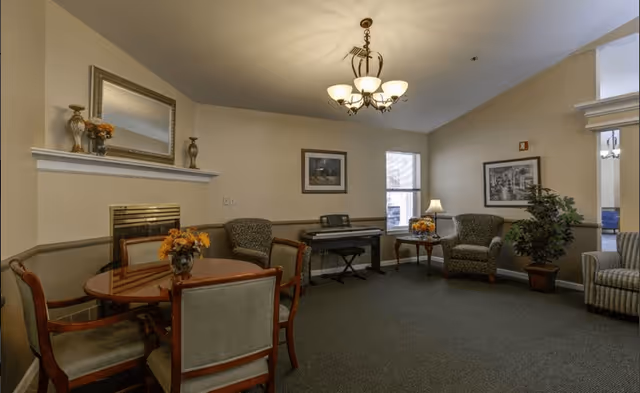 A cozy living room area with a round wooden table and four chairs, a fireplace with a mirror and decorative items on the mantel, two armchairs, a small side table with a lamp and flowers, a keyboard piano against the wall, framed pictures, a potted plant, and a chandelier hanging from the ceiling.