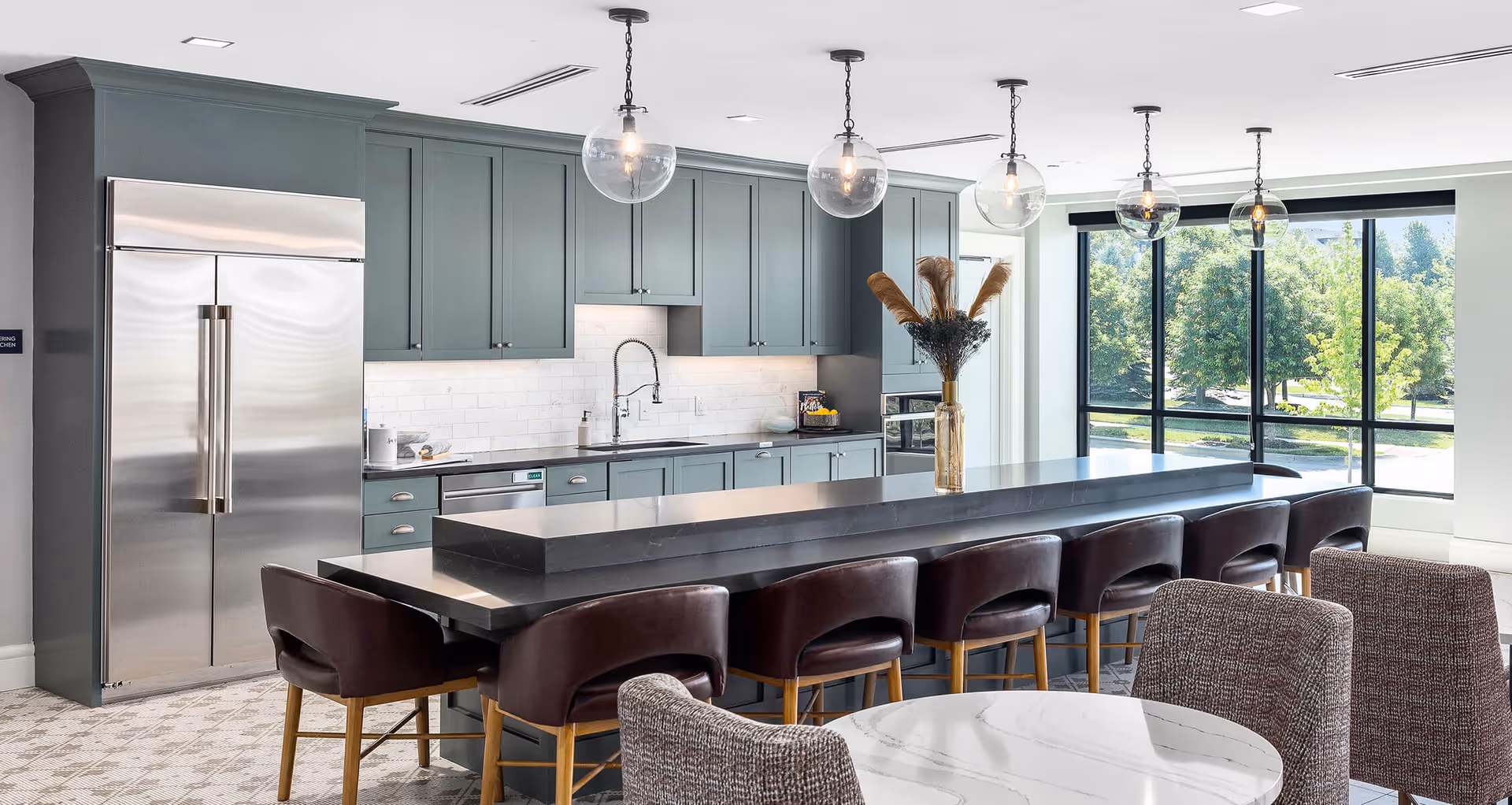Modern communal kitchen and dining area with a long dark island, leather bar stools, globe pendant lights, and large windows overlooking trees.