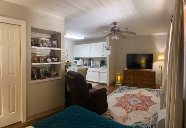 Interior view of a senior living facility room at Oakmont Estates showing a cozy living area with a brown recliner chair, a wooden TV stand with a flat-screen TV, a colorful patterned rug, and a ceiling fan. The room also includes a small kitchen area with white cabinets and appliances, and built-in shelves with framed photos and decorative items.