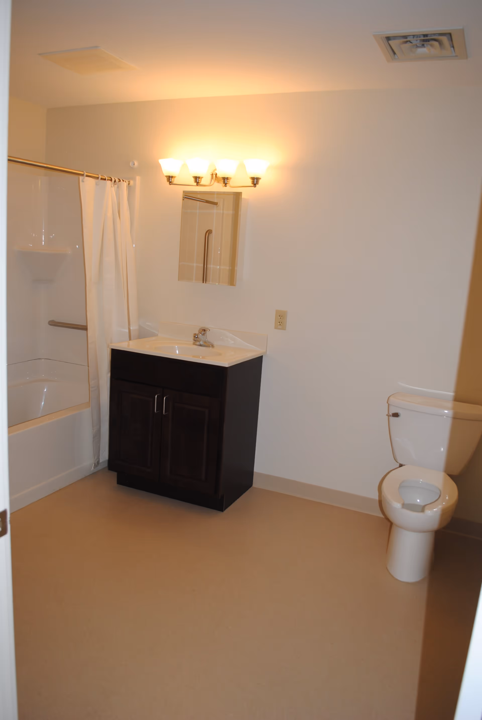 A simple bathroom with a vanity and mirror, a lit wall sconce, a shower with curtain, and a toilet.