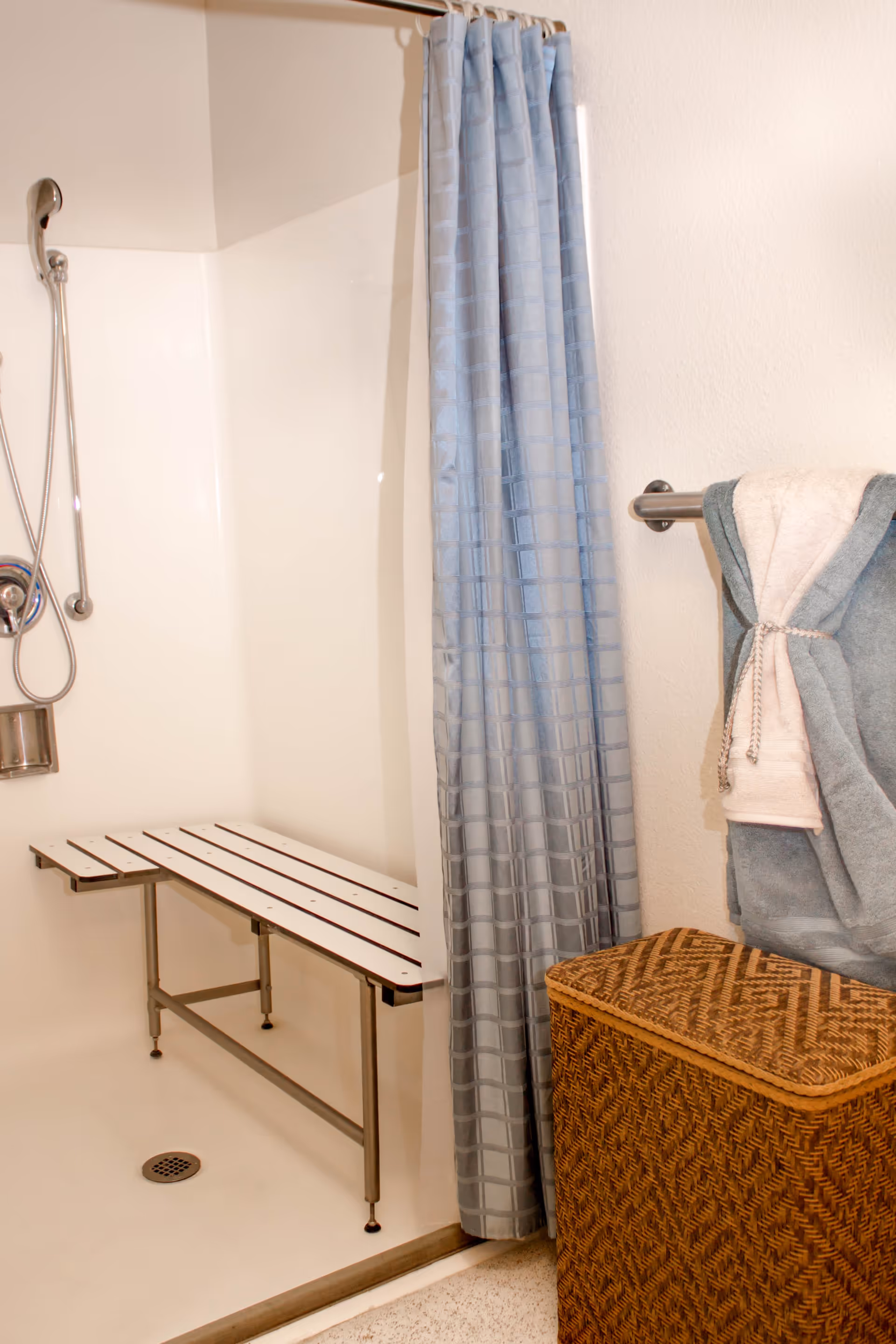 A bathroom shower area with a white foldable bench, a handheld showerhead, and a blue shower curtain. Next to the shower is a towel rack with folded towels and a woven laundry basket on the floor.
