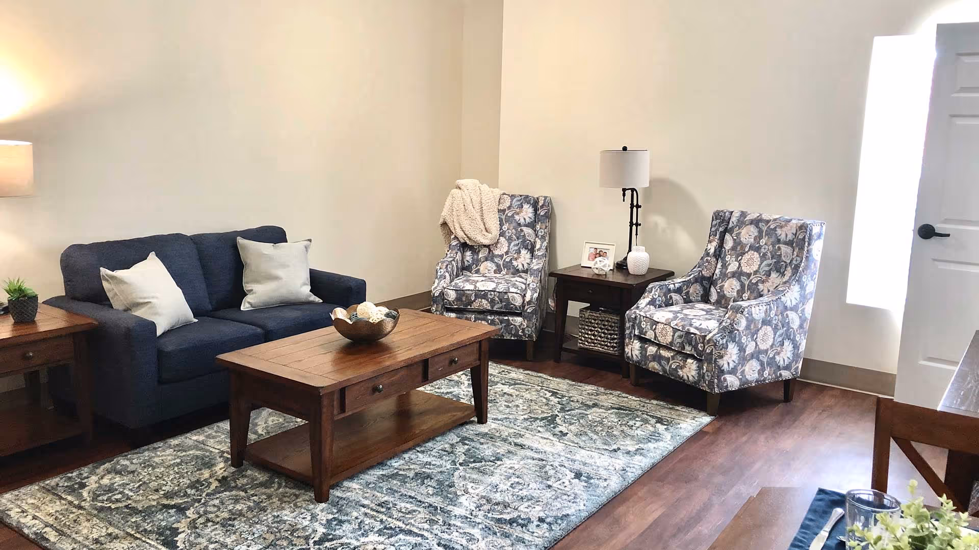 A cozy living room with a dark blue sofa adorned with two light-colored pillows, two floral patterned armchairs, a wooden coffee table with decorative items, a side table with a lamp and framed photo, and a patterned area rug on wooden flooring.