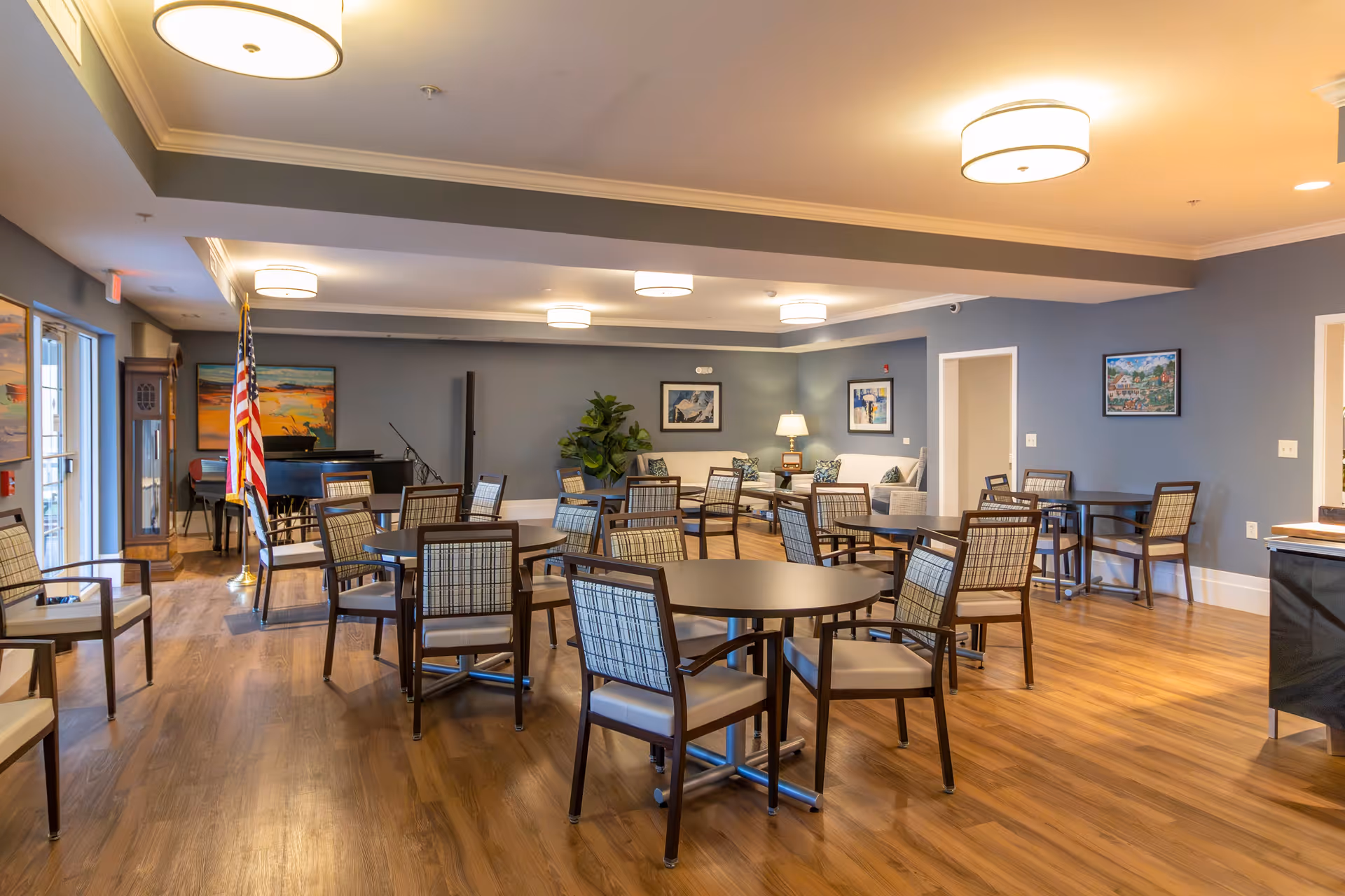 A spacious common area with multiple round tables and chairs arranged on a wooden floor. The room has blue-gray walls adorned with framed artwork and is well-lit with ceiling lights. There is a seating area with sofas and a lamp in the background, an American flag near a piano, and large windows on the left side letting in natural light.