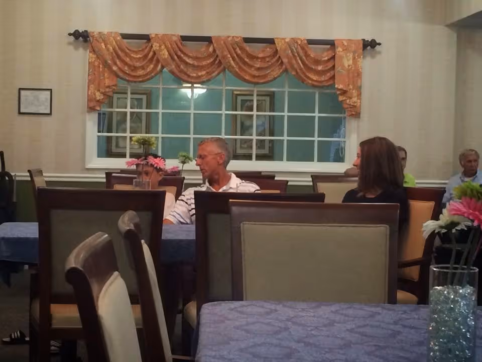 A dining room with several people seated at tables covered with blue tablecloths. The room has beige walls, a large window with orange floral valances, and framed pictures on the walls. There are flowers in vases on the tables.